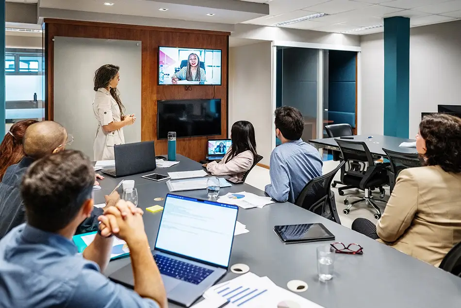 Multiple students sit around a large table while watching a television screen displaying someone on a video call.