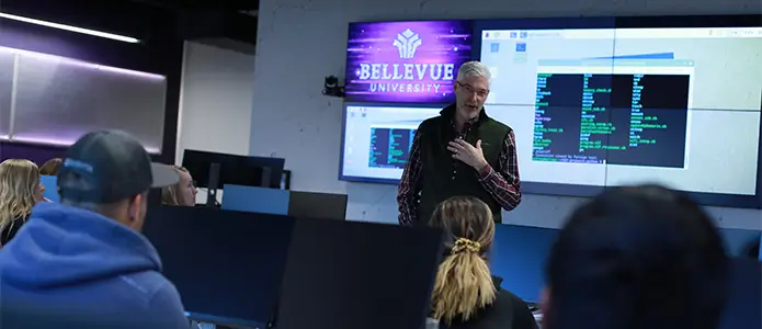 A professor stands in front of a wall-sized digital screen while instructing students in a computer lab.