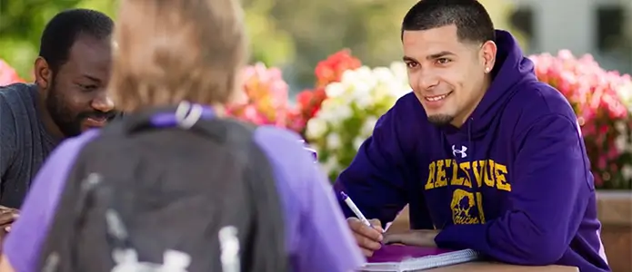 Multiple students wearing purple Bellevue University shirts work on coursework while seated at a table outside surrounded by flowers.