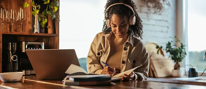 A student wearing headphones sits at a dining room table while taking notes and working on their laptop.> 