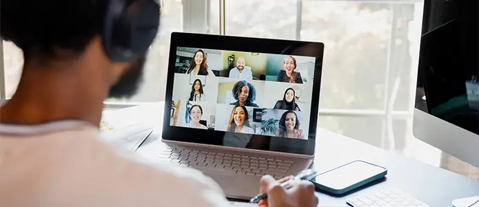 A student wearing headphones takes notes while engaging in an online video call with multiple other people.> 